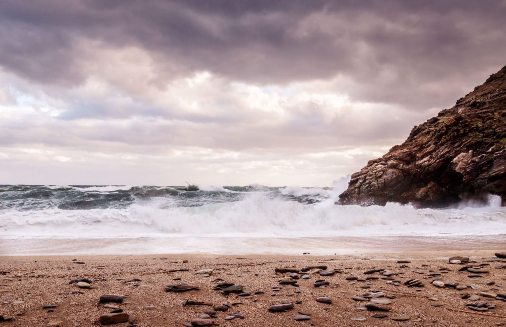 Big frothy waves crashing on a deserted, pepple beach in Greece. Jagged rocks and moody clouds in the sky create a dramatic scene.